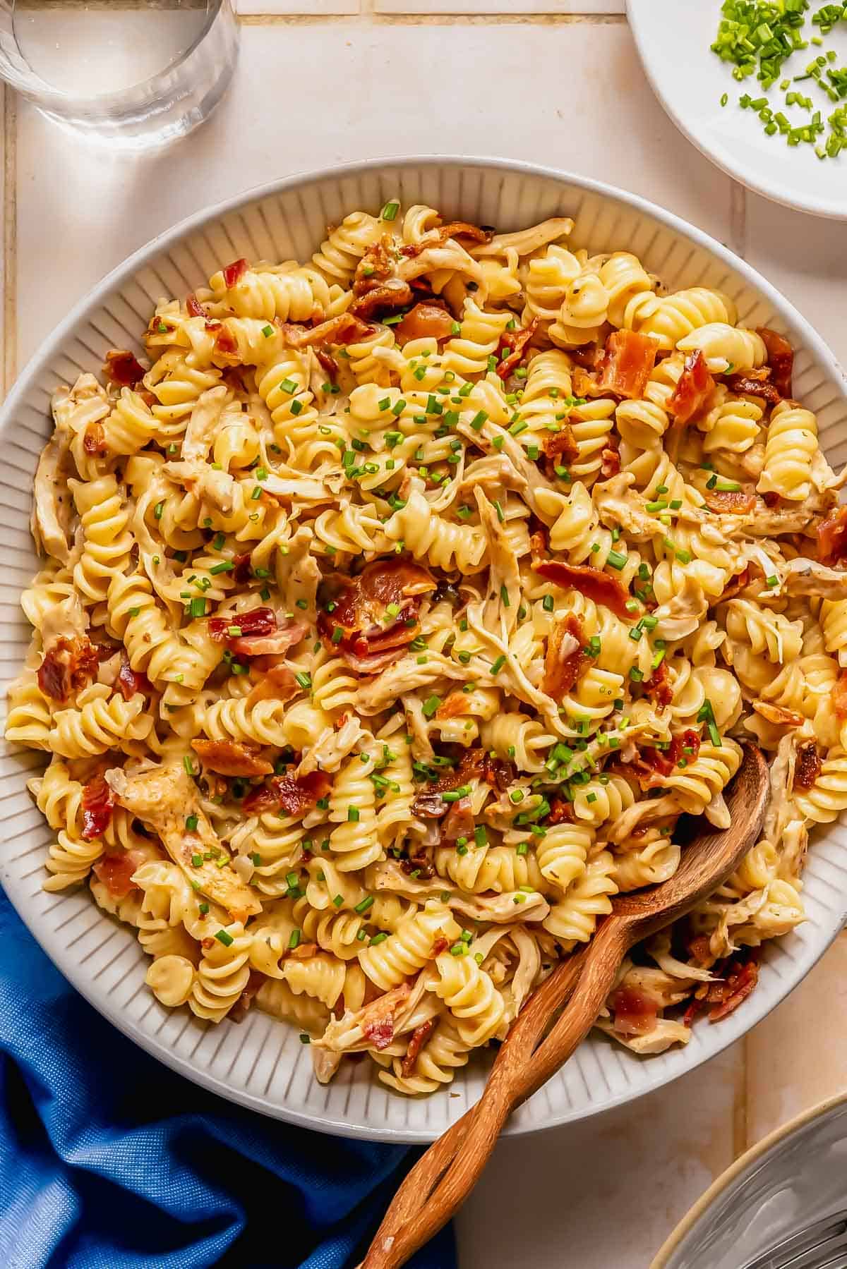 A plate of rotini pasta mixed with shredded chicken, crispy bacon, and chopped chives, with a wooden spoon resting in the dish.