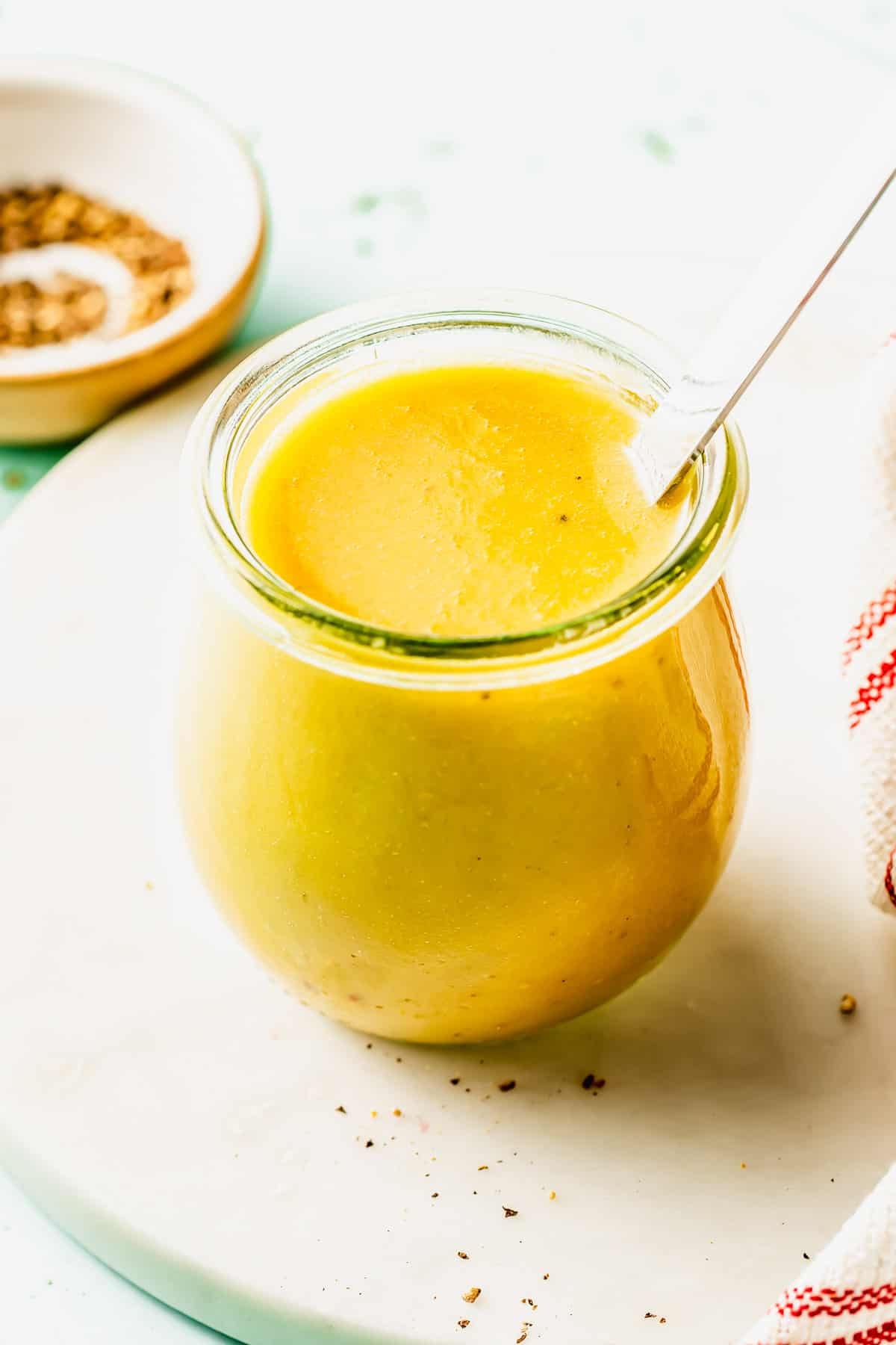 A glass jar filled with yellow liquid, possibly salad dressing or sauce, with a spoon inside. A small dish with spices and a striped cloth are nearby on a white surface.