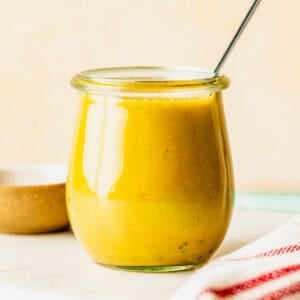 A small glass jar filled with yellow salad dressing, with a metal spoon inside, sits on a white surface next to a red striped towel.