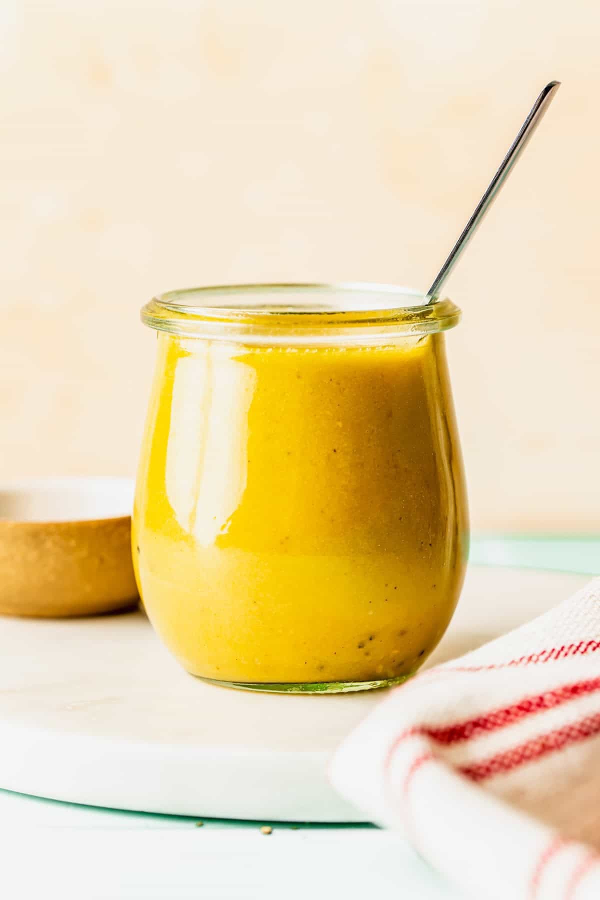 A small glass jar filled with yellow salad dressing, with a metal spoon inside, sits on a white surface next to a red striped towel.