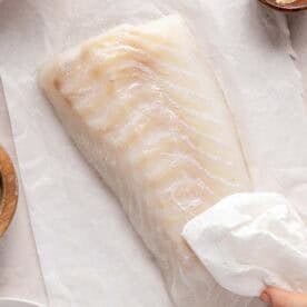 Raw haddock fillet on parchment paper with a hand patting it dry using a paper towel, surrounded by small bowls of ingredients.
