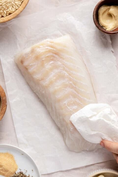 Raw haddock fillet on parchment paper with a hand patting it dry using a paper towel, surrounded by small bowls of ingredients.