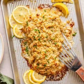 Baked crispy haddock fillet with a golden breadcrumb topping on a baking tray, garnished with parsley and lemon slices, with a spatula beside the fish.