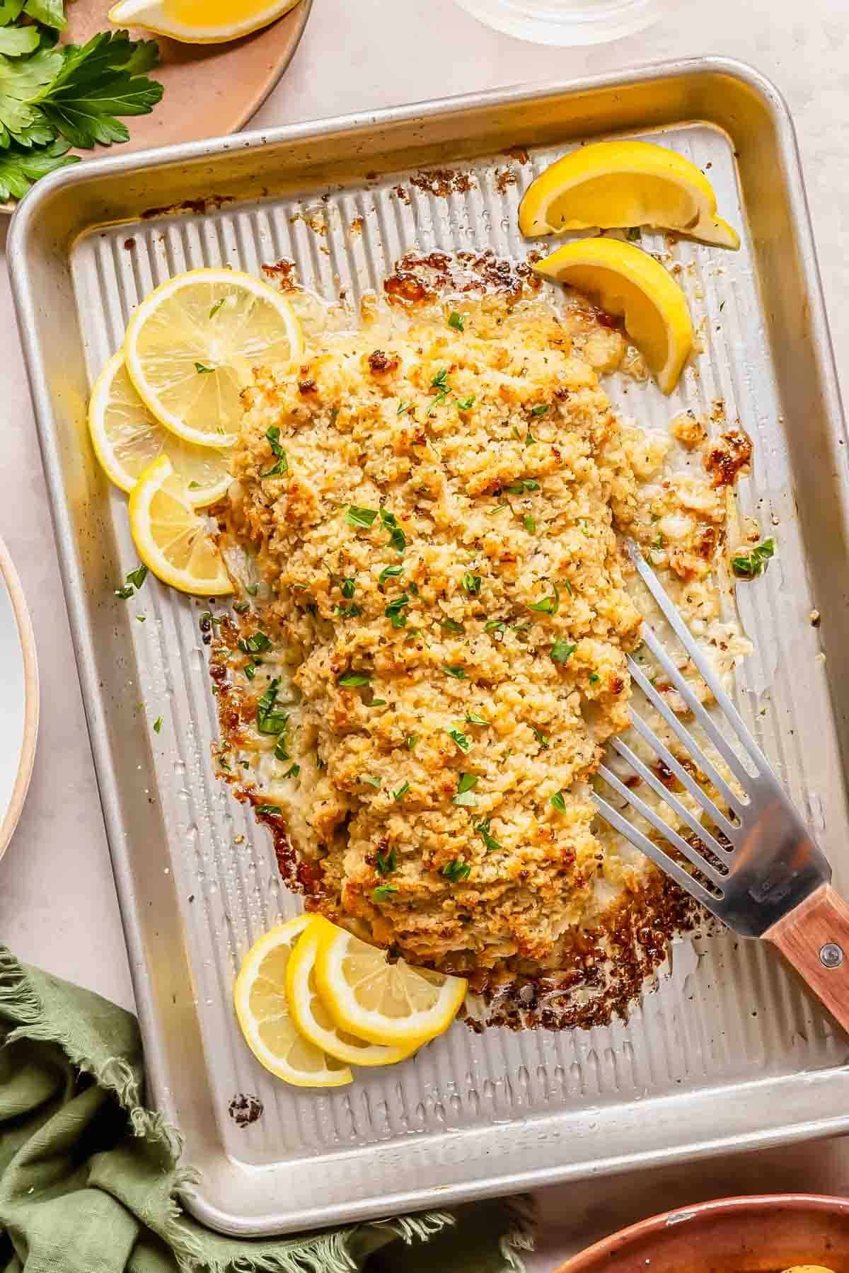 Baked crispy haddock fillet with a golden breadcrumb topping on a baking tray, garnished with parsley and lemon slices, with a spatula beside the fish.
