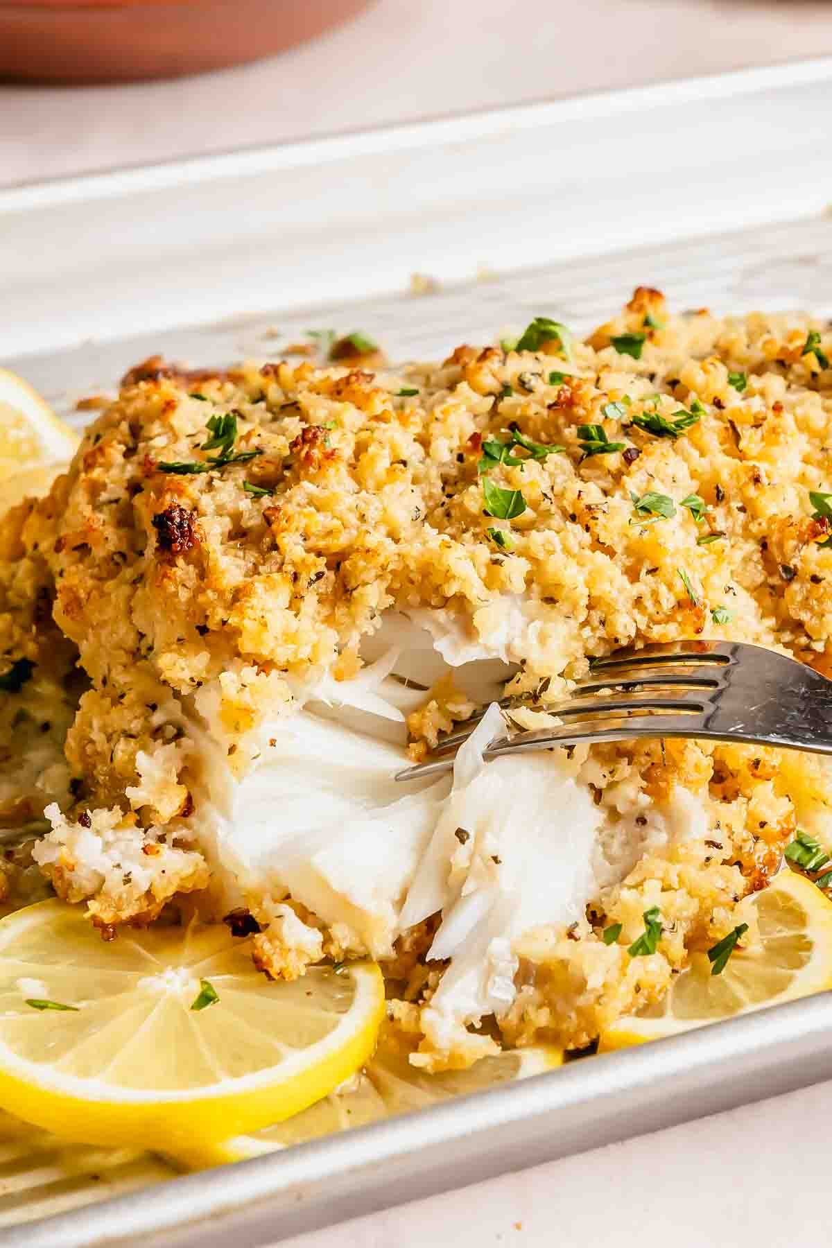 Baked crispy haddock with a golden breadcrumb crust, garnished with parsley and lemon slices, being flaked with a fork.