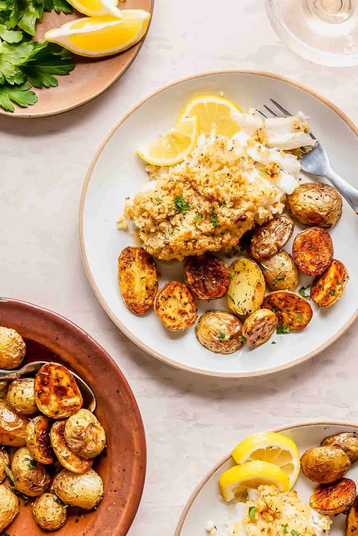 A plate with baked crispy haddock topped with breadcrumbs, roasted baby potatoes, lemon wedges, and fresh herbs, alongside dishes of extra potatoes and lemon slices.
