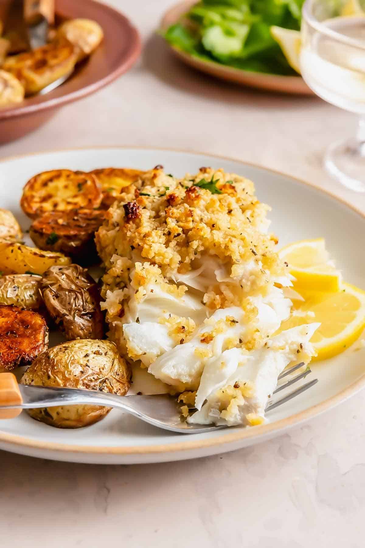 A plate with baked crispy haddock topped with crumbs, roasted baby potatoes, and lemon wedges, with a fork resting on the plate. A salad and drink are in the background.