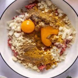 Ground meat, two raw egg yolks, chopped onions, breadcrumbs, and spices are arranged in a large white bowl, ready to be mixed for a recipe.