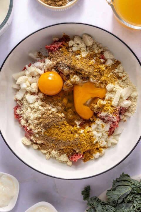 Ground meat, two raw egg yolks, chopped onions, breadcrumbs, and spices are arranged in a large white bowl, ready to be mixed for a recipe.