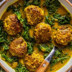A bowl of meatballs in a yellow curry sauce with kale, served with a wooden-handled spoon.