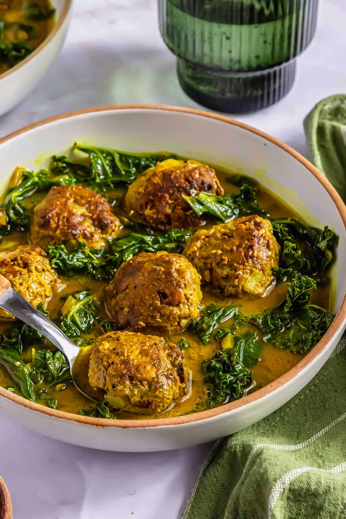 A bowl of meatballs and leafy greens in a curry broth, with a spoon resting in the bowl and a green glass in the background.