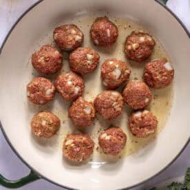 Raw meatballs with visible chopped onions are arranged in a large, white pot with a green rim. Some seasoning and leafy greens are nearby on the white surface.