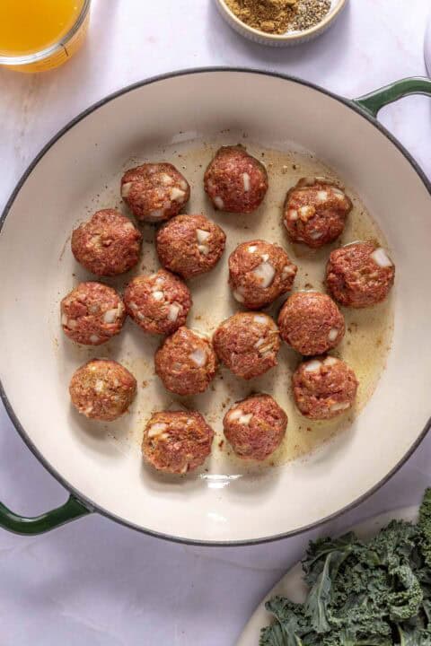 Raw meatballs with visible chopped onions are arranged in a large, white pot with a green rim. Some seasoning and leafy greens are nearby on the white surface.