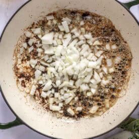 Chopped onions sizzling in a large white pan with oil, surrounded by bowls of broth, spices, and greens on a white surface.