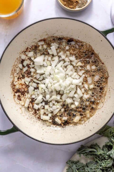 Chopped onions sizzling in a large white pan with oil, surrounded by bowls of broth, spices, and greens on a white surface.