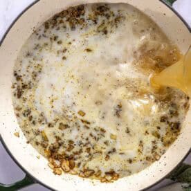 Broth is being poured into a pot with sautéed onions and spices, creating a bubbling mixture. Some leafy greens are visible on the side.