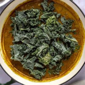 A white pot contains a yellow curry sauce topped with raw kale leaves. A white plate with food is partially visible in the upper left corner.