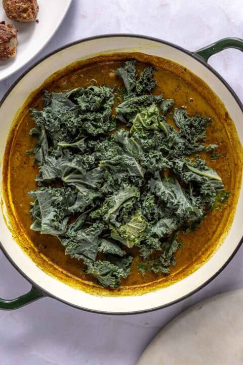 A white pot contains a yellow curry sauce topped with raw kale leaves. A white plate with food is partially visible in the upper left corner.