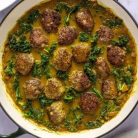 A white skillet filled with browned meatballs, leafy greens, and a yellow curry sauce, viewed from above on a light surface.