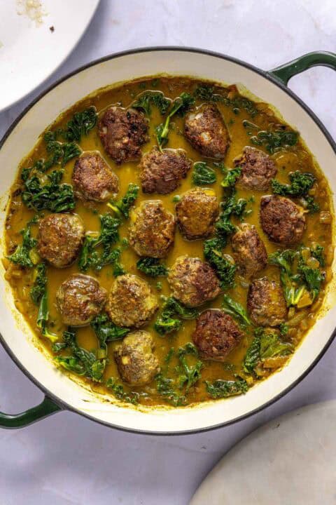 A white skillet filled with browned meatballs, leafy greens, and a yellow curry sauce, viewed from above on a light surface.