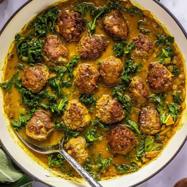 Meatballs and kale in a curry sauce are cooked in a large skillet, with a serving spoon resting inside. Nearby are pepper mills, a drinking glass, and a small bowl of seasoning.