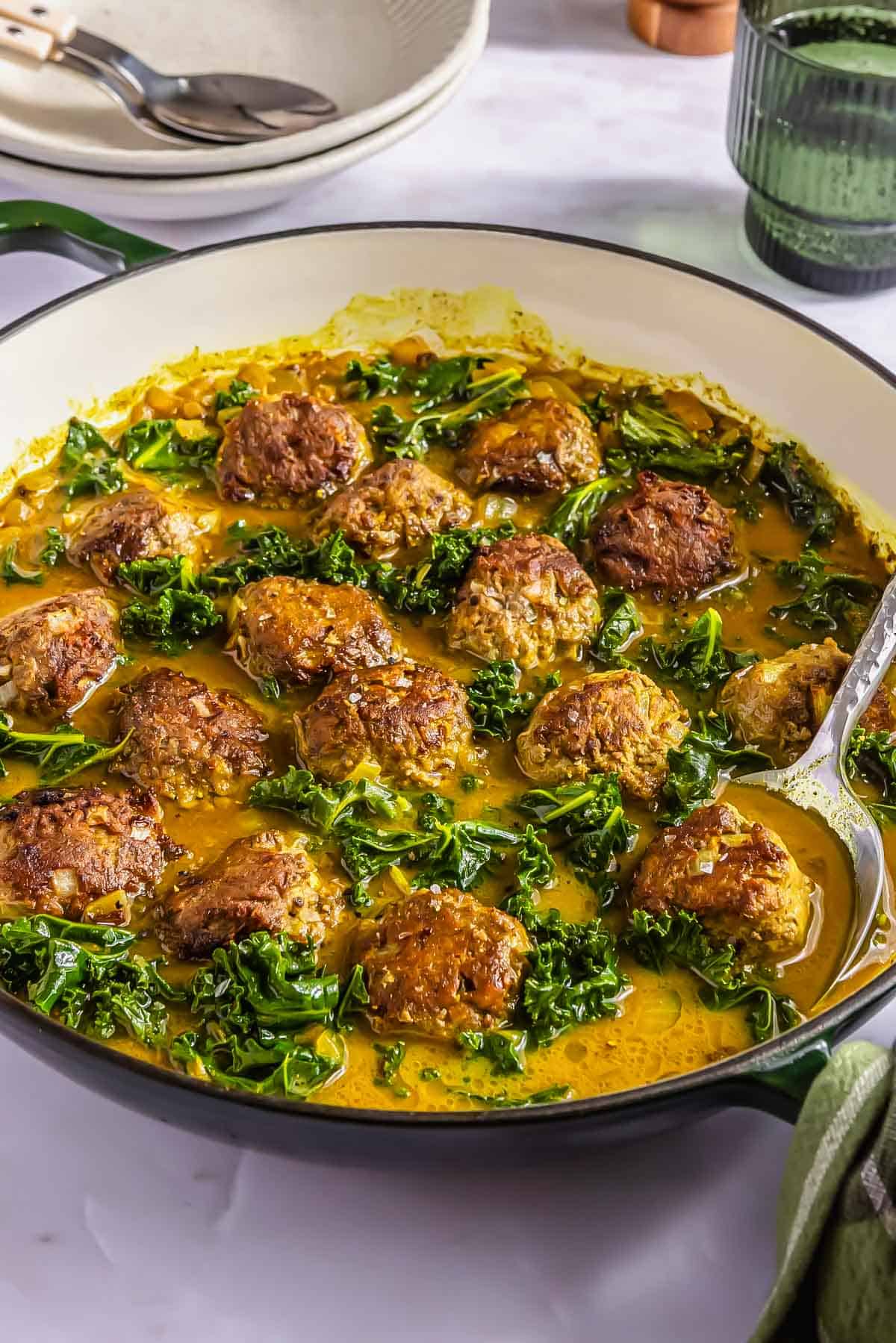 Meatballs cooked in a curry sauce with chopped kale in a large shallow pan, with a serving spoon resting on the side.
