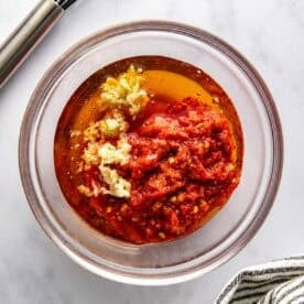 A glass bowl with harissa, minced garlic, olive oil, and spices on a marble surface, next to a whisk and a striped kitchen towel.