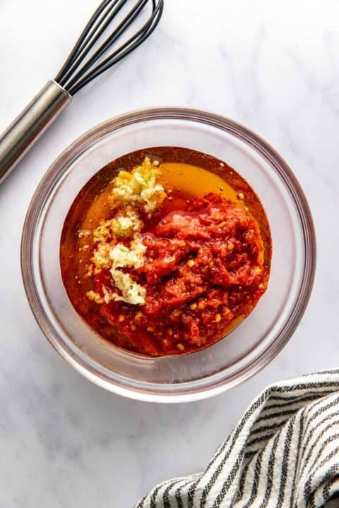 A glass bowl with harissa, minced garlic, olive oil, and spices on a marble surface, next to a whisk and a striped kitchen towel.