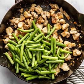 A cast iron skillet with cooked chicken pieces and fresh green beans, partially mixed, on a marble surface next to a striped towel.