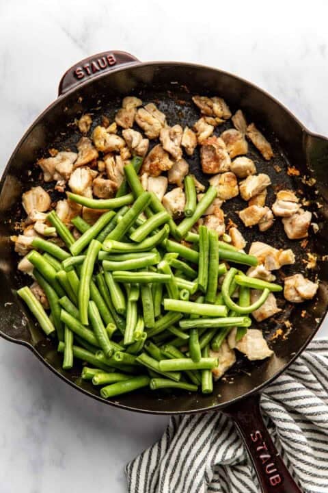 A cast iron skillet with cooked chicken pieces and fresh green beans, partially mixed, on a marble surface next to a striped towel.