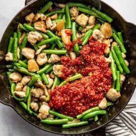 A cast iron pan with cooked chicken pieces, green beans, and a dollop of harissa-based sauce, next to a striped kitchen towel.
