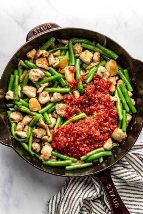 A cast iron pan with cooked chicken pieces, green beans, and a dollop of harissa-based sauce, next to a striped kitchen towel.