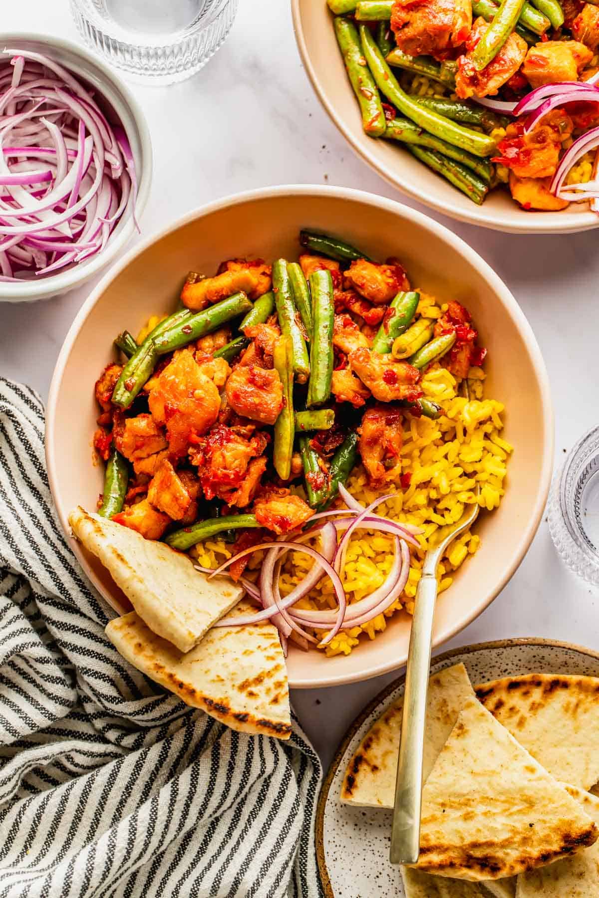 A bowl with yellow rice, sautéed chicken, green beans, red onions, and pita bread on the side. A bowl of red onions and a plate of pita wedges are nearby.