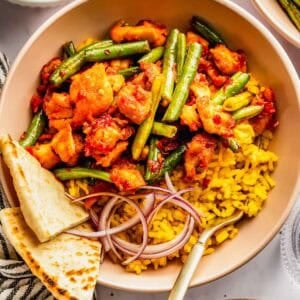 A bowl of yellow rice topped with green beans, spicy chicken, sliced red onions, and naan bread, with a fork resting inside.