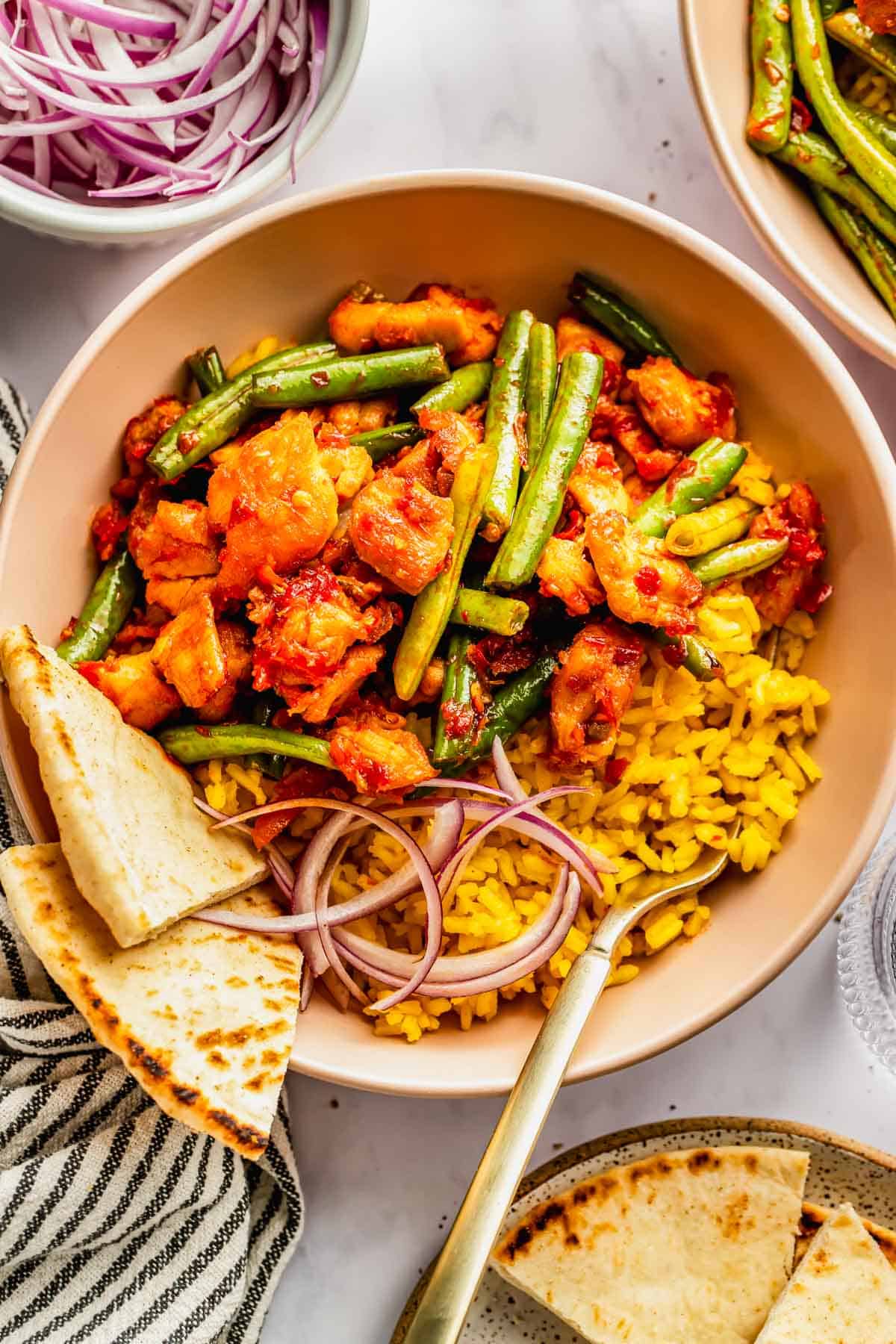 A bowl of yellow rice topped with green beans, spicy chicken, sliced red onions, and naan bread, with a fork resting inside.