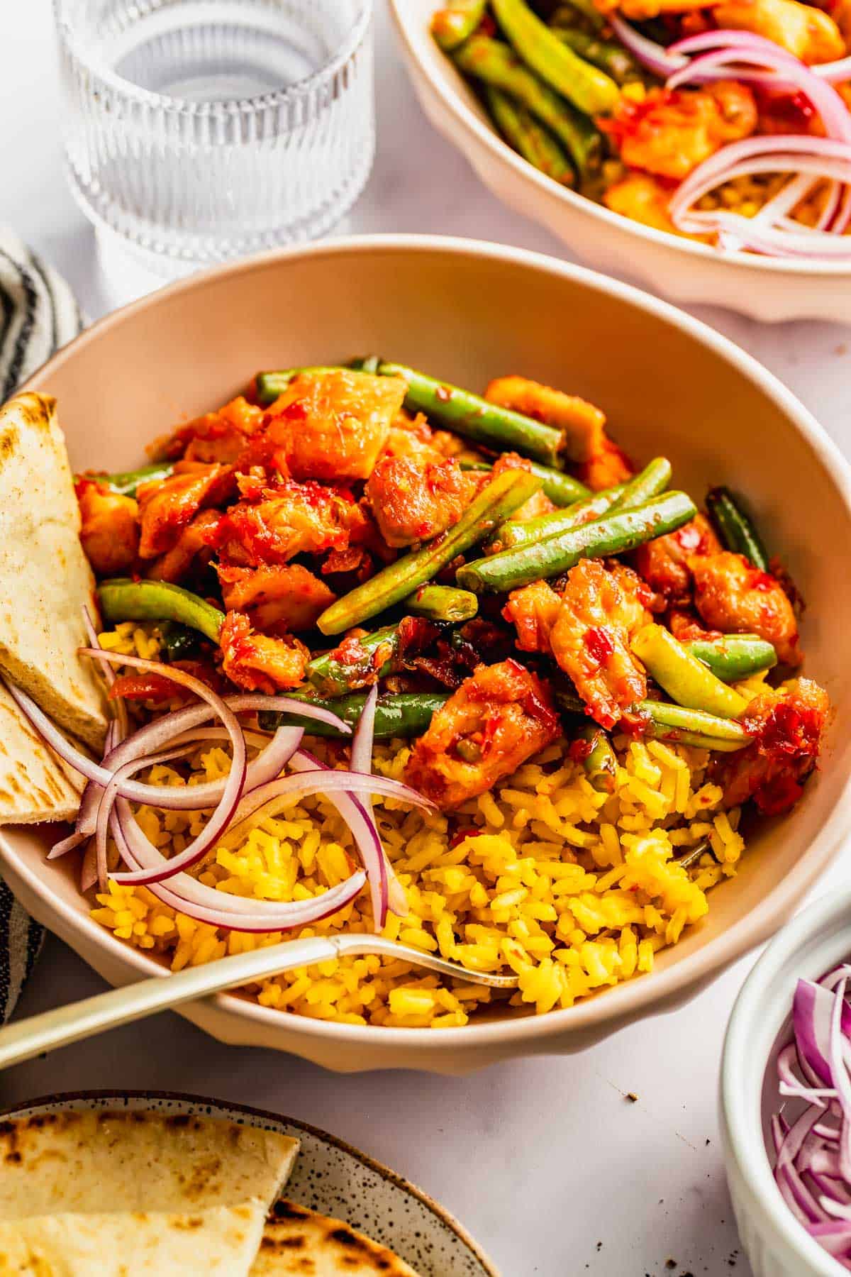 A bowl of yellow rice topped with saucy chicken, green beans, red onions, and flatbread, with a glass of water and another bowl in the background.
