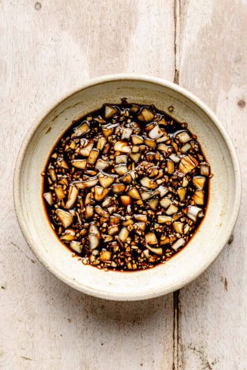 A bowl filled with chopped garlic mixed in a dark soy-based sauce, placed on a light wooden surface.