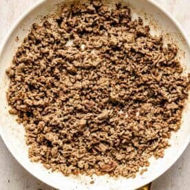 Cooked ground beef in a white skillet with a gold handle, sitting on a light wooden surface.