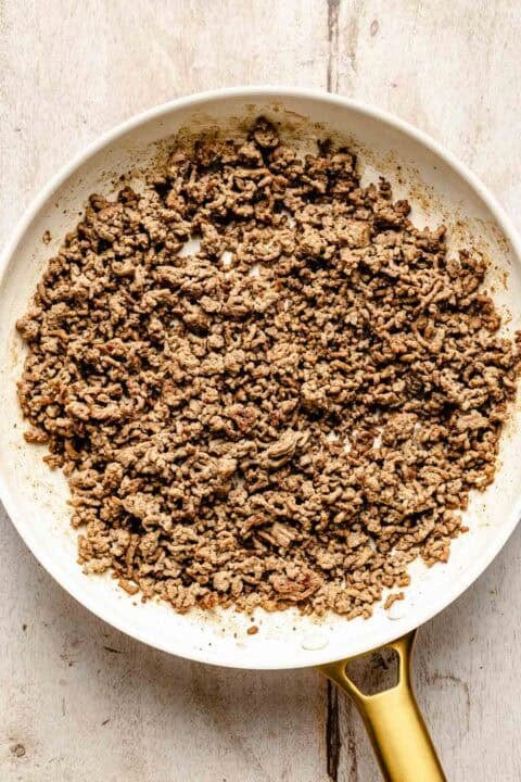 Cooked ground beef in a white skillet with a gold handle, sitting on a light wooden surface.