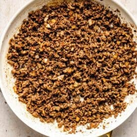 Cooked ground beef with diced onions in a white skillet, shown from above on a light wooden surface.