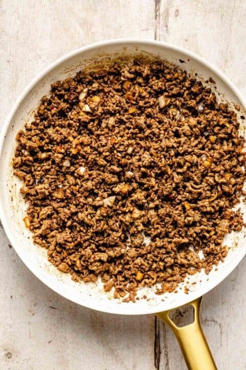 Cooked ground beef with diced onions in a white skillet, shown from above on a light wooden surface.