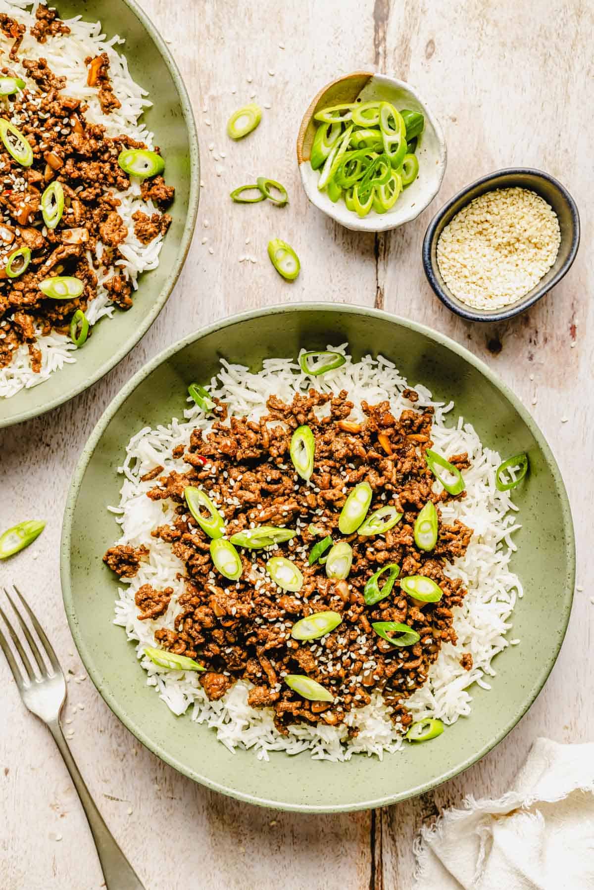 A green bowl filled with white rice topped with cooked ground meat, sliced green onions, and sesame seeds; small bowls of sliced onions and sesame seeds nearby.