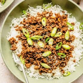 A bowl of white rice topped with cooked ground beef, sliced green onions, and sesame seeds, with a fork on the side.