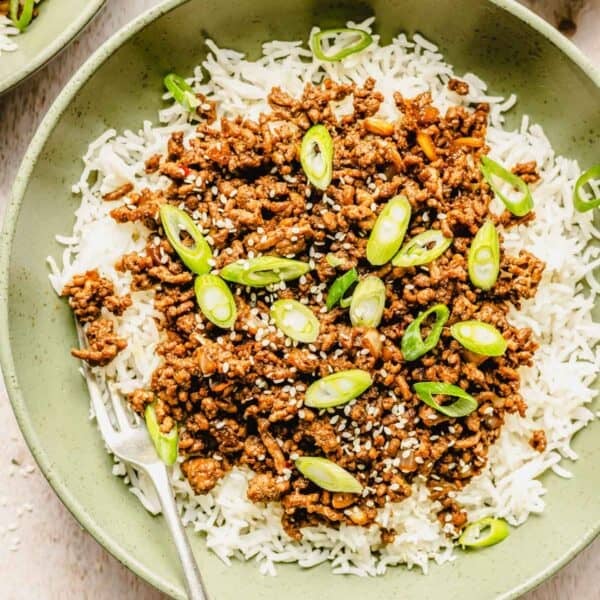 A bowl of white rice topped with cooked ground beef, sliced green onions, and sesame seeds, with a fork on the side.
