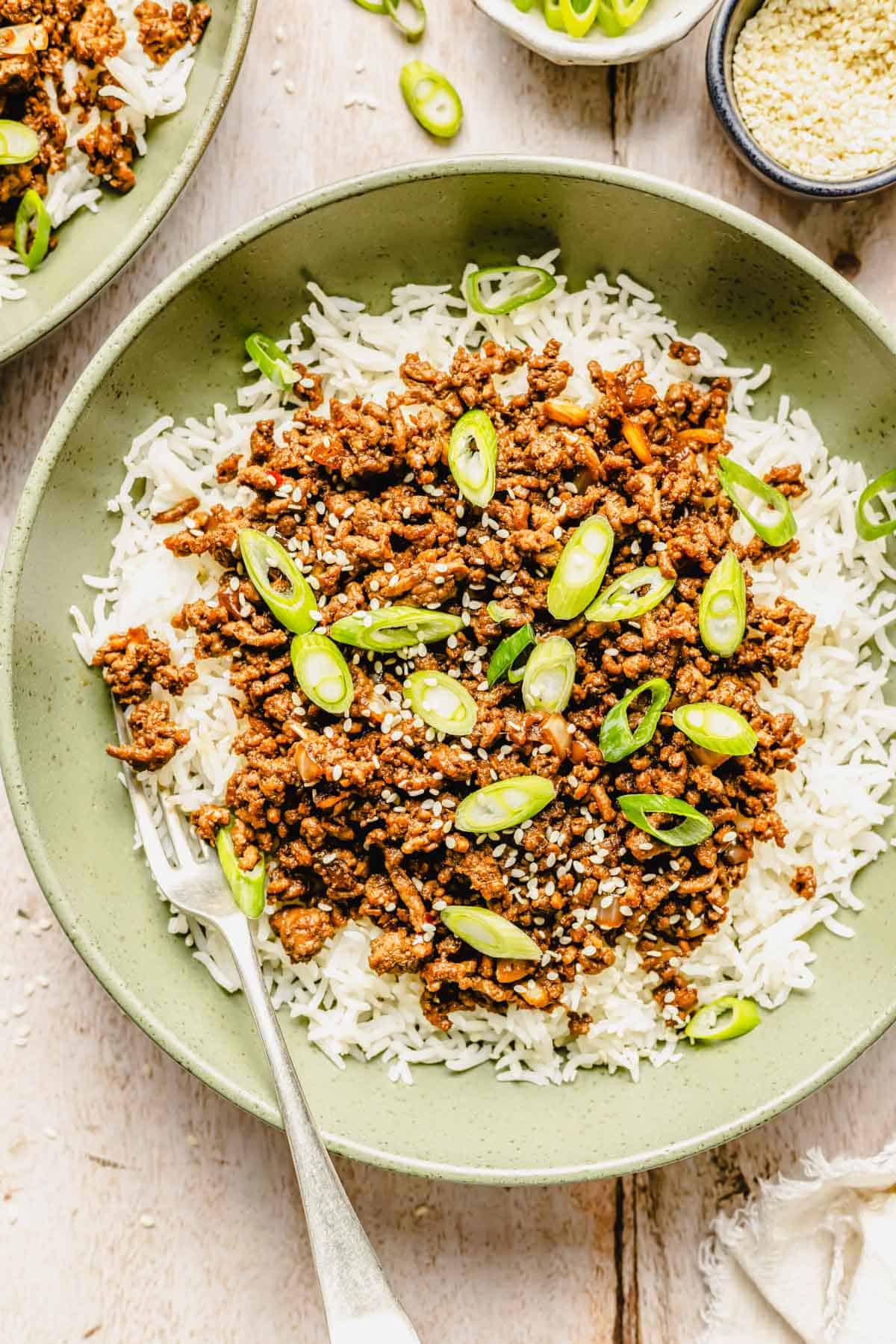 A bowl of white rice topped with cooked ground beef, sliced green onions, and sesame seeds, with a fork on the side.