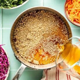 A measuring cup pours broth into a saucepan with quinoa, surrounded by bowls of chopped kale, celery, carrots, and cabbage on a tiled countertop.