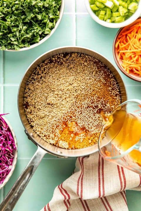 A measuring cup pours broth into a saucepan with quinoa, surrounded by bowls of chopped kale, celery, carrots, and cabbage on a tiled countertop.