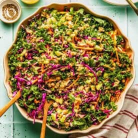 Large bowl of chopped kale salad with shredded carrots, red cabbage, green onions, peanuts, and dressing, placed on a light surface with utensils and a striped napkin nearby.