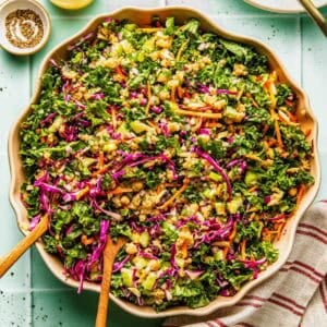 Large bowl of chopped kale salad with shredded carrots, red cabbage, green onions, peanuts, and dressing, placed on a light surface with utensils and a striped napkin nearby.