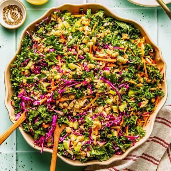 Large bowl of chopped kale salad with shredded carrots, red cabbage, green onions, peanuts, and dressing, placed on a light surface with utensils and a striped napkin nearby.